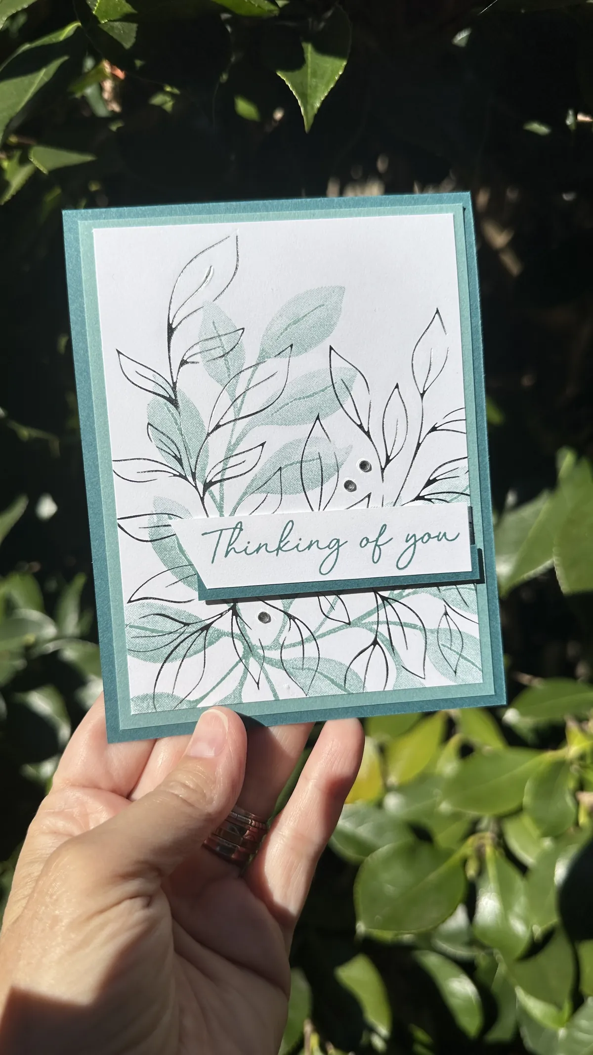 Thinking of You card with silver hot foil leaves and Lost Lagoon stamped leaves on a Pretty Peacock card base, held in front of green foliage